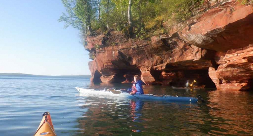 a student paddles a kayak beside a tall red cliff